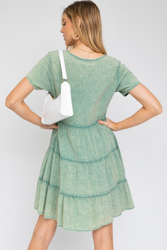 A woman in a Washed Baby Doll Mini Dress, featuring a vintage-inspired green design with a tiered skirt, holds a white handbag, perfectly poised for casual outings against a plain background.