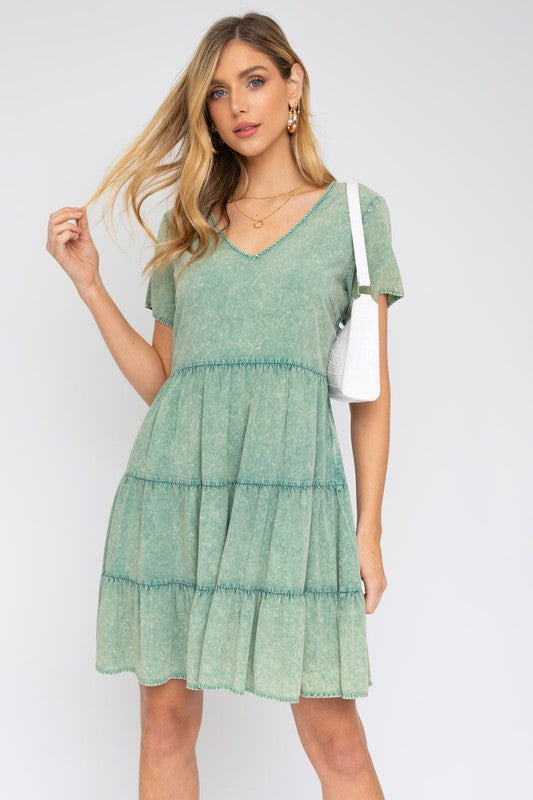 A woman in a Washed Baby Doll Mini Dress, featuring a vintage-inspired green design with a tiered skirt, holds a white handbag, perfectly poised for casual outings against a plain background.