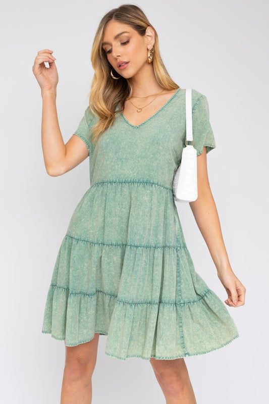 A woman in a Washed Baby Doll Mini Dress, featuring a vintage-inspired green design with a tiered skirt, holds a white handbag, perfectly poised for casual outings against a plain background.