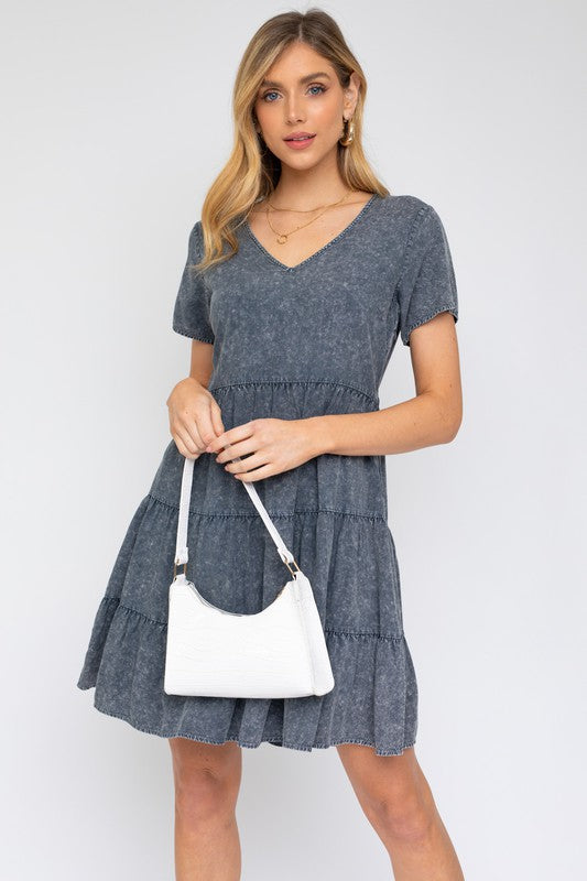 A woman in a Washed Baby Doll Mini Dress, featuring a vintage-inspired green design with a tiered skirt, holds a white handbag, perfectly poised for casual outings against a plain background.