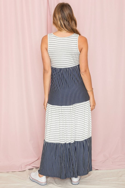 A person stands against a pink backdrop wearing the Plus Sleeveless Stripe Tiered Maxi Dress, made from a comfortable rayon spandex blend with alternating white and navy blue stripes, paired with white sneakers.