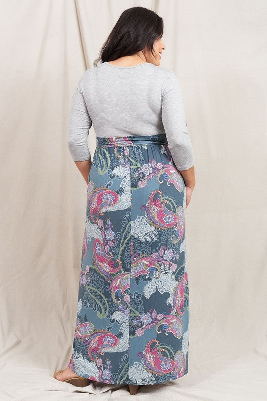 A woman in a Plus Sash Maxi Dress with Pockets, featuring long sleeves in gray and a long, colorful paisley-patterned skirt, stands against a beige backdrop.