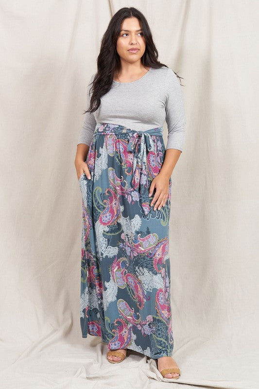 A woman in a Plus Sash Maxi Dress with Pockets, featuring long sleeves in gray and a long, colorful paisley-patterned skirt, stands against a beige backdrop.