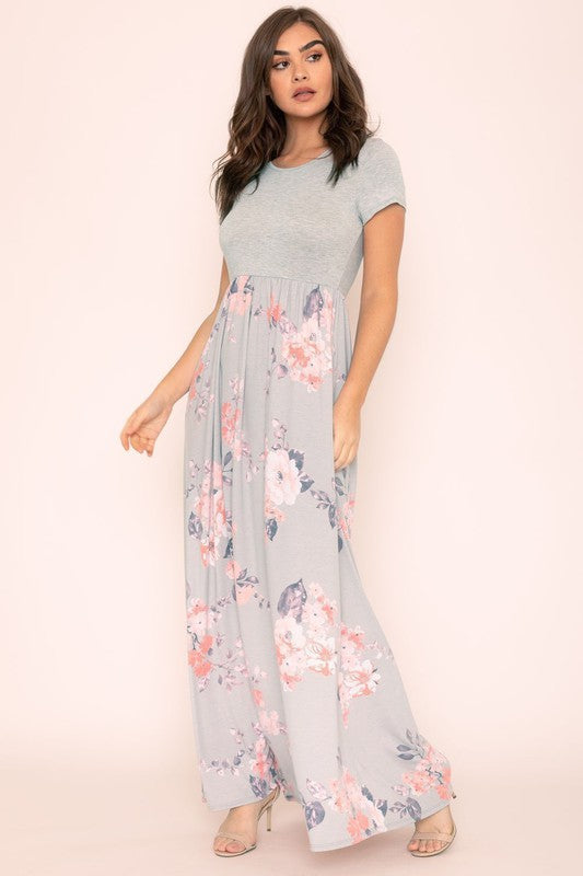 A woman wearing the Plus Short Sleeve Floral Maxi Dress stands smiling, her hands slightly above her waist, against a plain background. Made in the United States, her attire exudes charm and elegance.