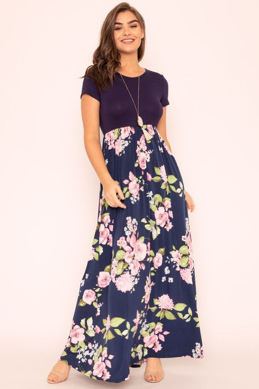 A woman wearing the Plus Short Sleeve Floral Maxi Dress stands smiling, her hands slightly above her waist, against a plain background. Made in the United States, her attire exudes charm and elegance.