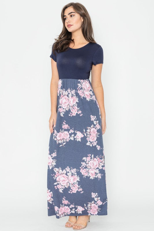 A woman wearing the Plus Short Sleeve Floral Maxi Dress stands smiling, her hands slightly above her waist, against a plain background. Made in the United States, her attire exudes charm and elegance.