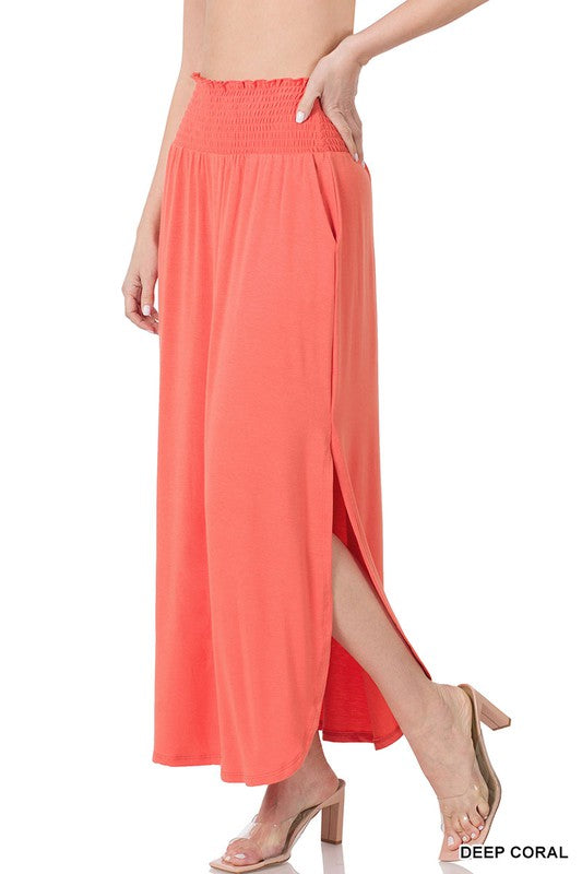 A person is elegantly dressed in a Smocked Waist Side Slit Maxi Skirt with Pockets in deep coral, beautifully paired with clear heeled sandals.