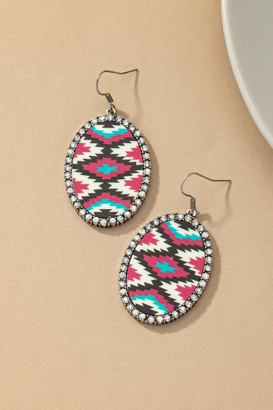 The Aztec oval drop earrings with rhinestones feature two acrylic drops that showcase bold geometric Aztec designs in black, white, red, and teal. Each earring is beautifully accented with a sparkling glass rhinestone border set against a beige background.