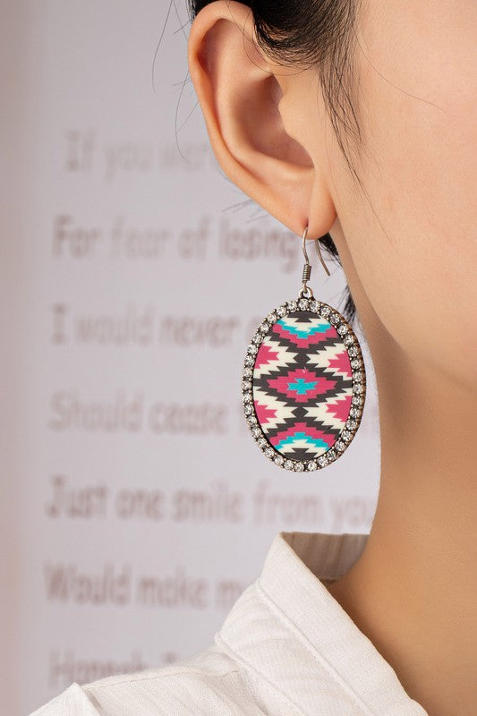 The Aztec oval drop earrings with rhinestones feature two acrylic drops that showcase bold geometric Aztec designs in black, white, red, and teal. Each earring is beautifully accented with a sparkling glass rhinestone border set against a beige background.