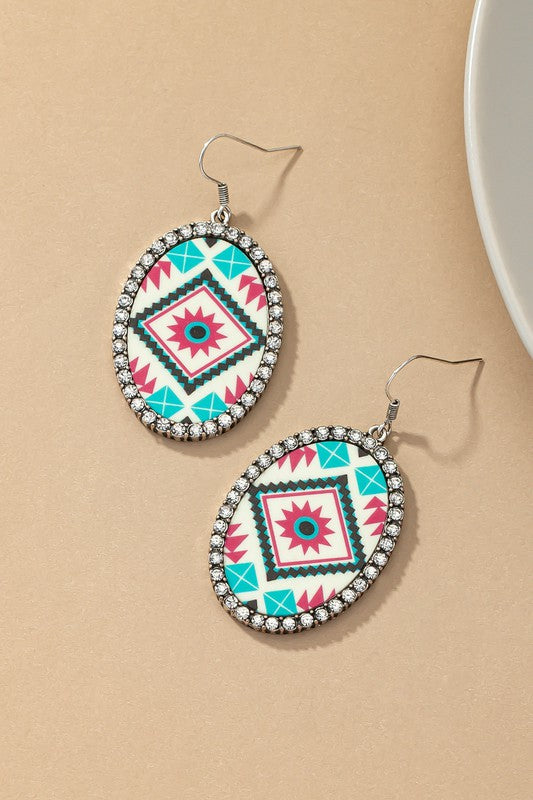 A pair of Aztec oval drop earrings with rhinestones, featuring colorful geometric patterns and bordered by glass rhinestones, is placed on a tan surface near a white dish.