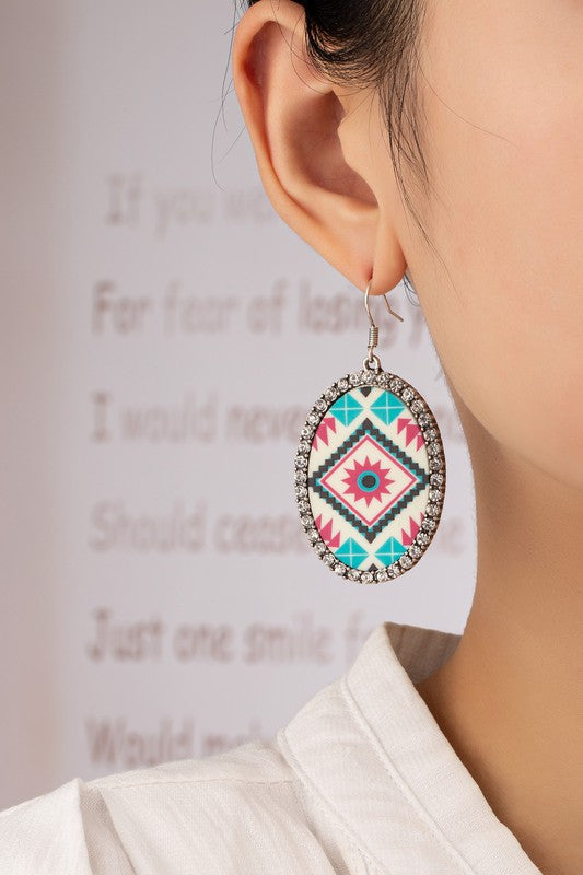 A pair of Aztec oval drop earrings with rhinestones, featuring colorful geometric patterns and bordered by glass rhinestones, is placed on a tan surface near a white dish.