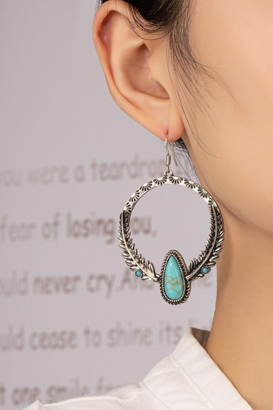 A pair of Boho distressed feather circle earrings in silver, adorned with turquoise teardrop-shaped stones and featuring intricate feather-like detailing, are displayed on a light surface.