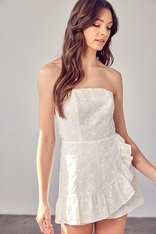 A woman in an Eyelet Ruffle Romper with strapless design and ruffle accents stands against a light background, her long and wavy hair cascading around her shoulders.