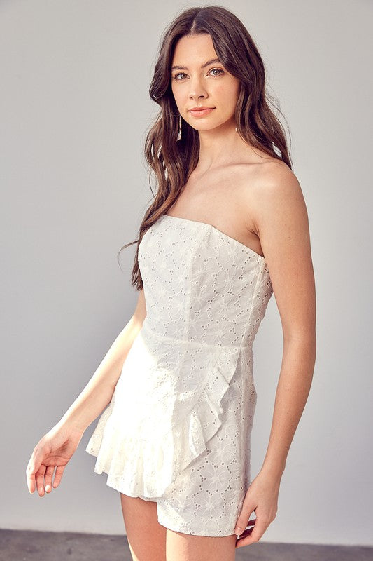 A woman in an Eyelet Ruffle Romper with strapless design and ruffle accents stands against a light background, her long and wavy hair cascading around her shoulders.