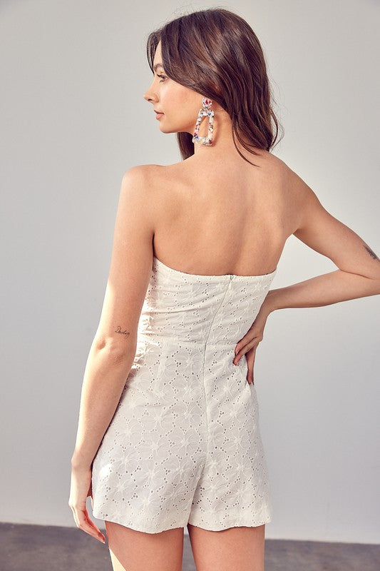 A woman in an Eyelet Ruffle Romper with strapless design and ruffle accents stands against a light background, her long and wavy hair cascading around her shoulders.