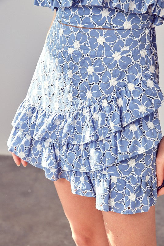 A person wearing the Eyelet Ruffle Skirt in blue, featuring white floral patterns and ruffled layers, makes an elegant wardrobe addition.