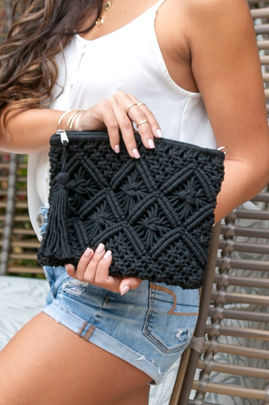 A person sits with the Macrame Tassel Clutch, which showcases a geometric pattern and tassel, secured with a zipper closure. They wear a white top and denim shorts that perfectly complement this stylish accessory.