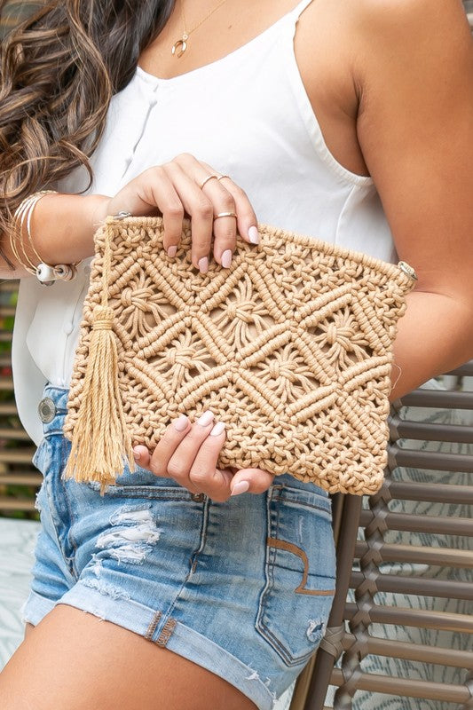 A person sits with the Macrame Tassel Clutch, which showcases a geometric pattern and tassel, secured with a zipper closure. They wear a white top and denim shorts that perfectly complement this stylish accessory.