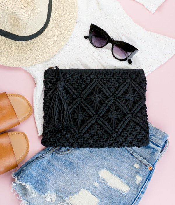 A person sits with the Macrame Tassel Clutch, which showcases a geometric pattern and tassel, secured with a zipper closure. They wear a white top and denim shorts that perfectly complement this stylish accessory.