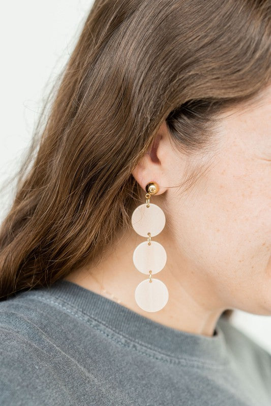 The Elle Earrings - Linen are a pair of dangling earrings featuring three circular beige pendants on each side, crafted from lightweight acrylic with a subtle rose gold shimmer, artfully displayed on a white rectangular dish set on a wooden surface.