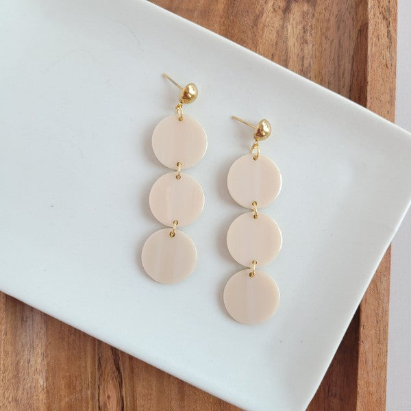 The Elle Earrings - Linen are a pair of dangling earrings featuring three circular beige pendants on each side, crafted from lightweight acrylic with a subtle rose gold shimmer, artfully displayed on a white rectangular dish set on a wooden surface.