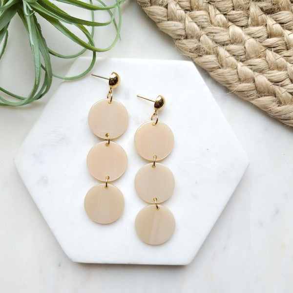 The Elle Earrings - Linen are a pair of dangling earrings featuring three circular beige pendants on each side, crafted from lightweight acrylic with a subtle rose gold shimmer, artfully displayed on a white rectangular dish set on a wooden surface.