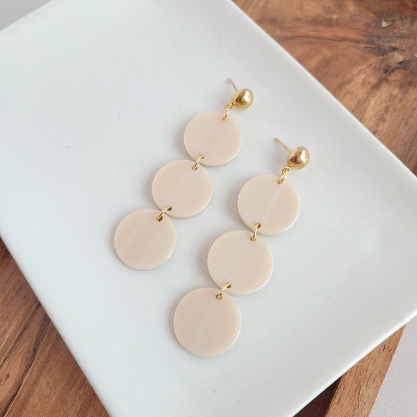 The Elle Earrings - Linen are a pair of dangling earrings featuring three circular beige pendants on each side, crafted from lightweight acrylic with a subtle rose gold shimmer, artfully displayed on a white rectangular dish set on a wooden surface.