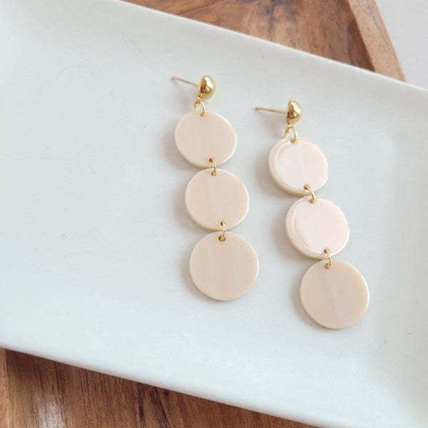 The Elle Earrings - Linen are a pair of dangling earrings featuring three circular beige pendants on each side, crafted from lightweight acrylic with a subtle rose gold shimmer, artfully displayed on a white rectangular dish set on a wooden surface.