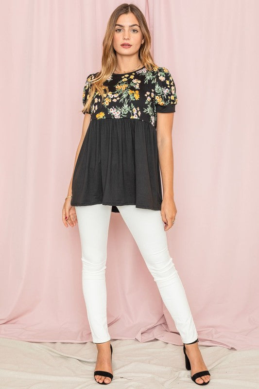 The woman stands confidently in the Plus Baby Doll Floral Puff Sleeve Tunic. Its charming puff sleeves accentuate the floral print, perfectly complementing the navy bottom half as she poses against a light pink curtain.