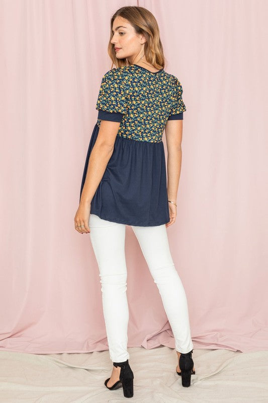 The woman stands confidently in the Plus Baby Doll Floral Puff Sleeve Tunic. Its charming puff sleeves accentuate the floral print, perfectly complementing the navy bottom half as she poses against a light pink curtain.