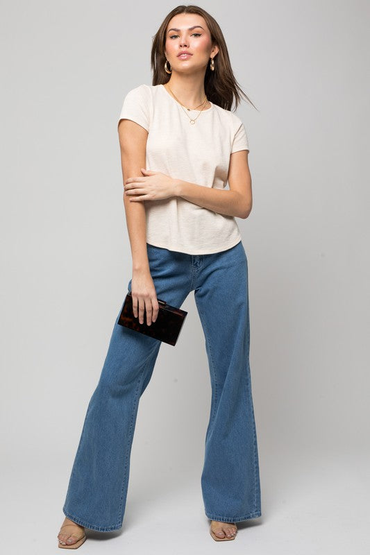 A woman wearing the S/S Back Button Down Rib Top and blue jeans is holding a small bag while smiling against a neutral background.