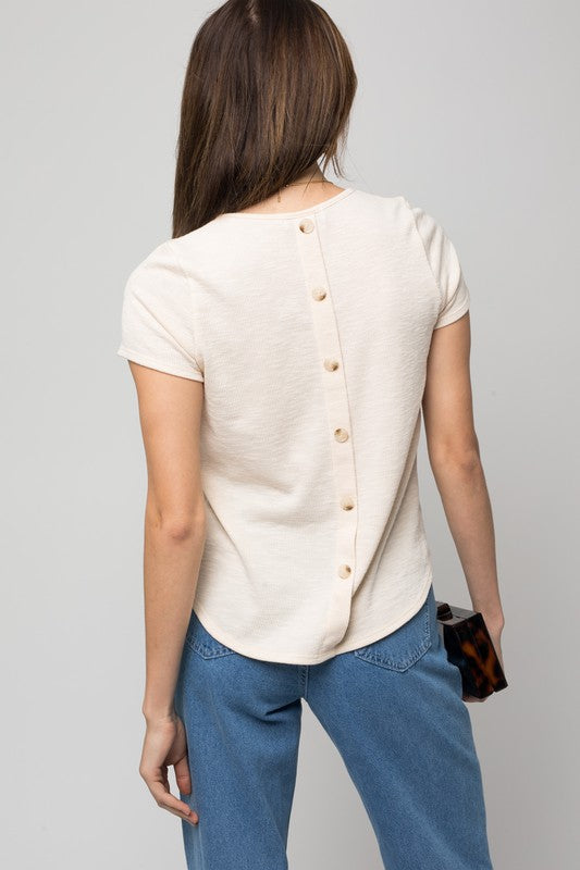 A woman wearing the S/S Back Button Down Rib Top and blue jeans is holding a small bag while smiling against a neutral background.