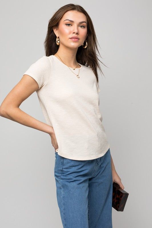 A woman wearing the S/S Back Button Down Rib Top and blue jeans is holding a small bag while smiling against a neutral background.