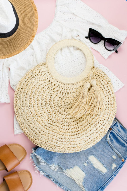 A woman holding a Straw Tassel O-Ring Tote, wearing a white sleeveless top, denim shorts, and a bracelet.