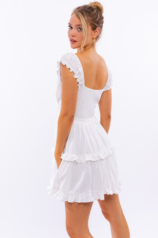 A person is kneeling in the Cap Sleeve Ruffle Mini Dress, which showcases exquisite ruffle detailing and floral lace trim, set against a white background decorated with pink star-like patterns.