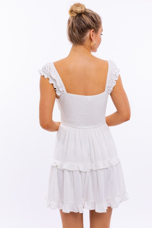 A person is kneeling in the Cap Sleeve Ruffle Mini Dress, which showcases exquisite ruffle detailing and floral lace trim, set against a white background decorated with pink star-like patterns.