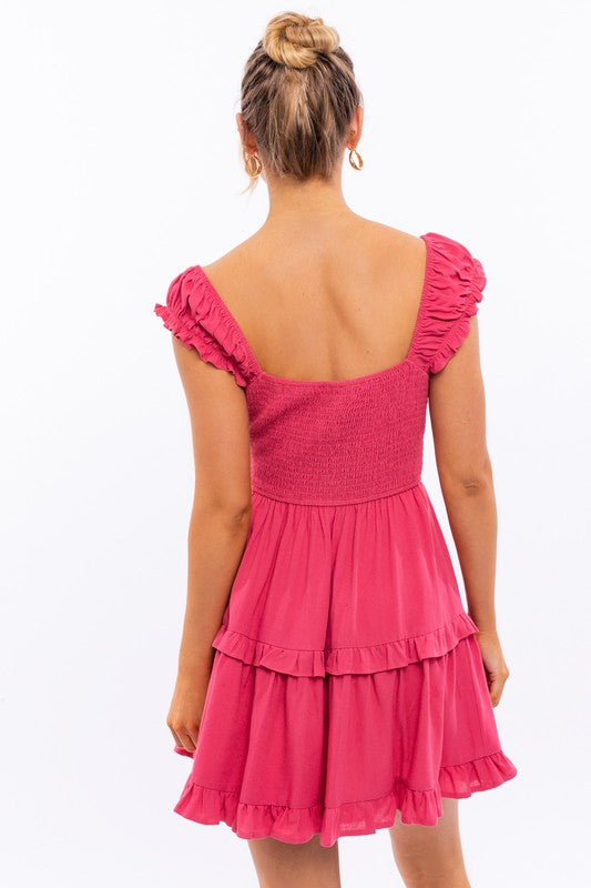 A person is kneeling in the Cap Sleeve Ruffle Mini Dress, which showcases exquisite ruffle detailing and floral lace trim, set against a white background decorated with pink star-like patterns.