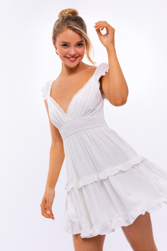 A person is kneeling in the Cap Sleeve Ruffle Mini Dress, which showcases exquisite ruffle detailing and floral lace trim, set against a white background decorated with pink star-like patterns.