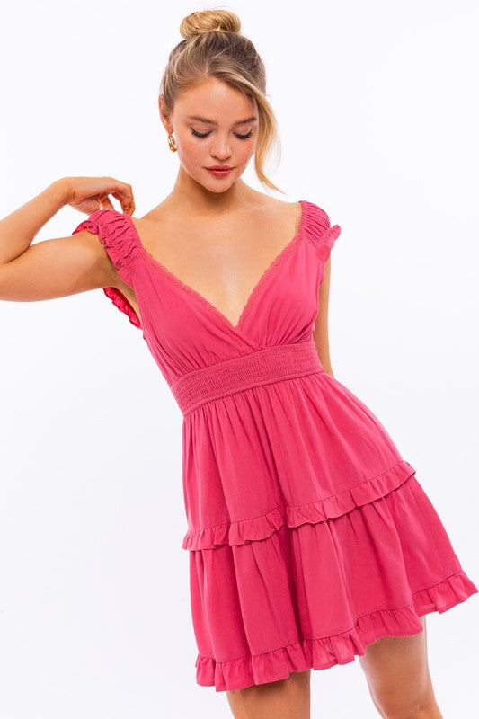 A person is kneeling in the Cap Sleeve Ruffle Mini Dress, which showcases exquisite ruffle detailing and floral lace trim, set against a white background decorated with pink star-like patterns.