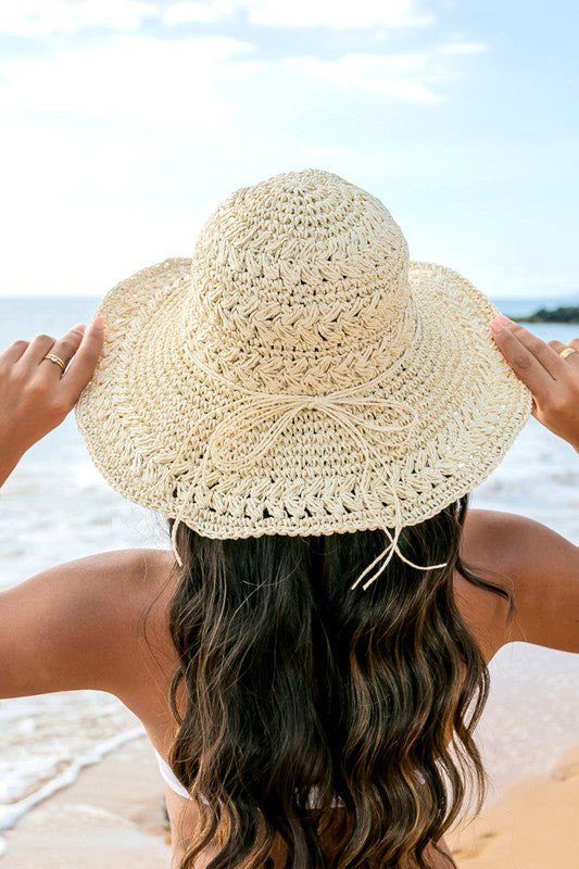 A woman in a white swimsuit smiles while holding onto her Woven Straw Sunhat with a raffia bow at the beach.