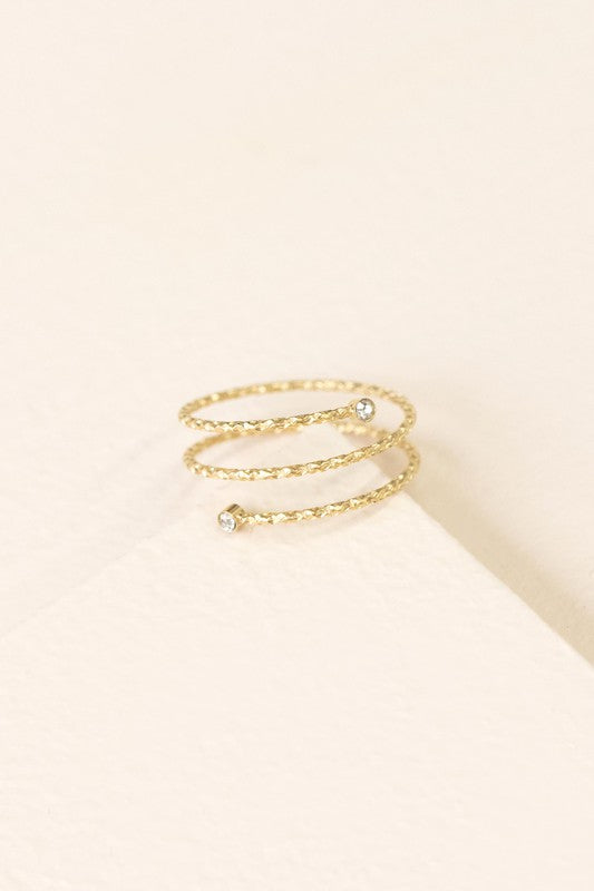 The Round and Round Ring boasts a stunning gold spiral design embellished with two small diamond accents, crafted from stainless steel with 14k gold plating, elegantly presented on a light beige background.