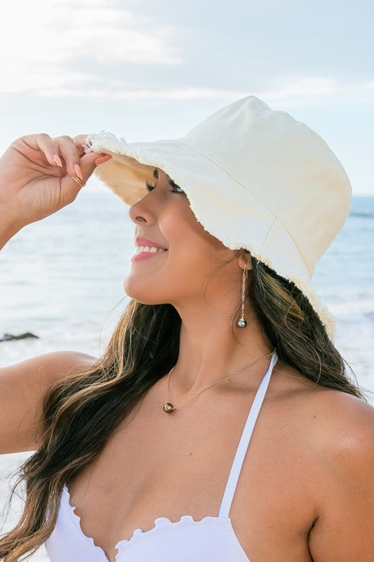 Woman in a white swimsuit and Frayed Edge Canvas Bucket Hat at the beach looking into the distance.