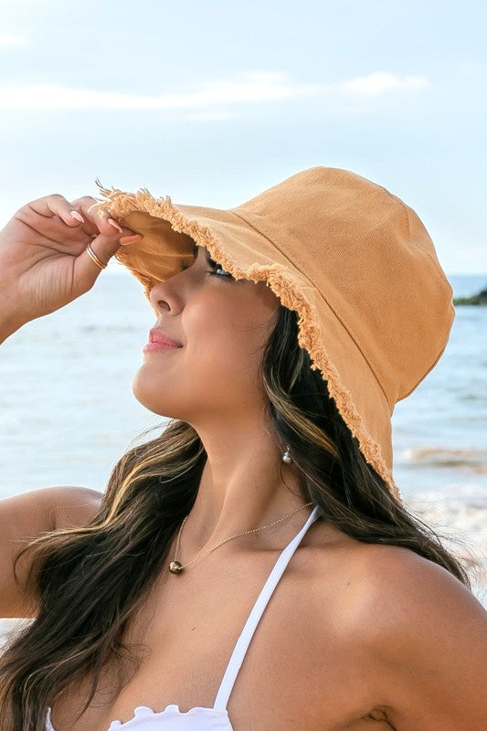 Woman in a white swimsuit and Frayed Edge Canvas Bucket Hat at the beach looking into the distance.