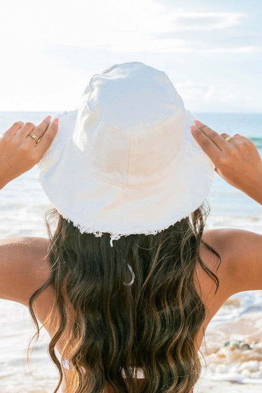 Woman in a white swimsuit and Frayed Edge Canvas Bucket Hat at the beach looking into the distance.