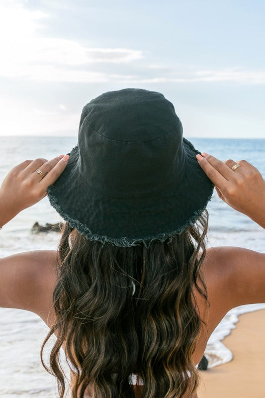Woman in a white swimsuit and Frayed Edge Canvas Bucket Hat at the beach looking into the distance.