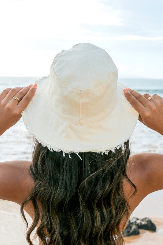 Woman in a white swimsuit and Frayed Edge Canvas Bucket Hat at the beach looking into the distance.