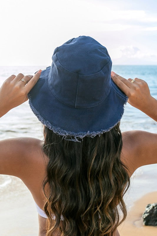Woman in a white swimsuit and Frayed Edge Canvas Bucket Hat at the beach looking into the distance.