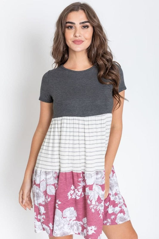 A woman dons the Short Sleeve Floral Tiered Midi Dress, showcasing a dark top, striped middle section, and a floral-patterned skirt. Made in the USA, this elegant dress creates a striking silhouette against its plain backdrop.