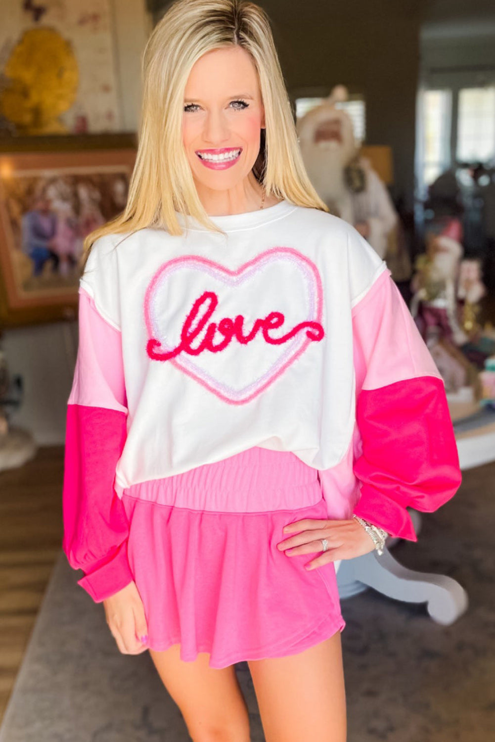 A woman with long blonde hair is smiling and posing indoors. She is wearing a White Tinsel Love Heart Graphic Color Block Sleeve Sweatshirt with pink shorts. There are framed photos and a table in the background.