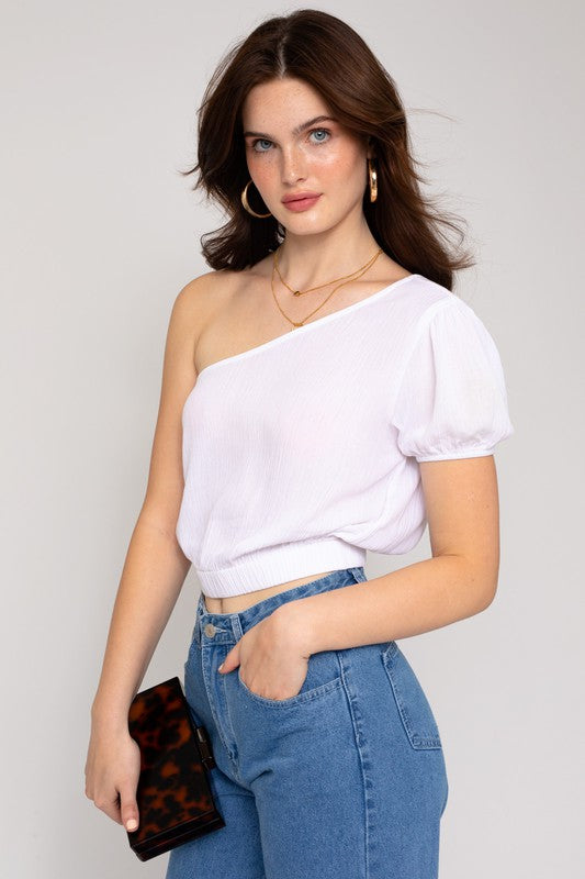 A person in a fashionable wardrobe featuring an asymmetrical design, wearing the S/S One Shoulder Top and jeans, holds a small rectangular purse while posing against a plain background.